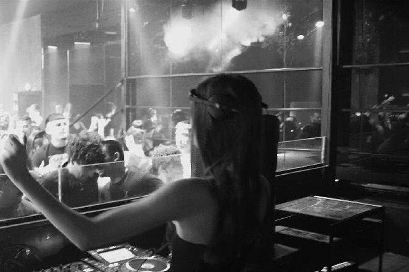 Carla Ros, DJ de hard techno, pinchando en cabina frente a la pista, fotografía en blanco y negro · PLAYNOW