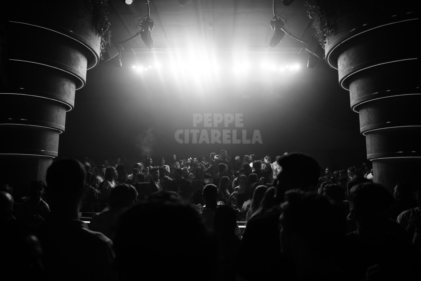 Sala llena durante una noche de Peppe Citarella, con su nombre proyectado sobre el escenario, en blanco y negro · PLAYNOW
